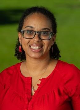 Woman wearing glasses and wearing red earrings and red blouse, standing in front of greenery.